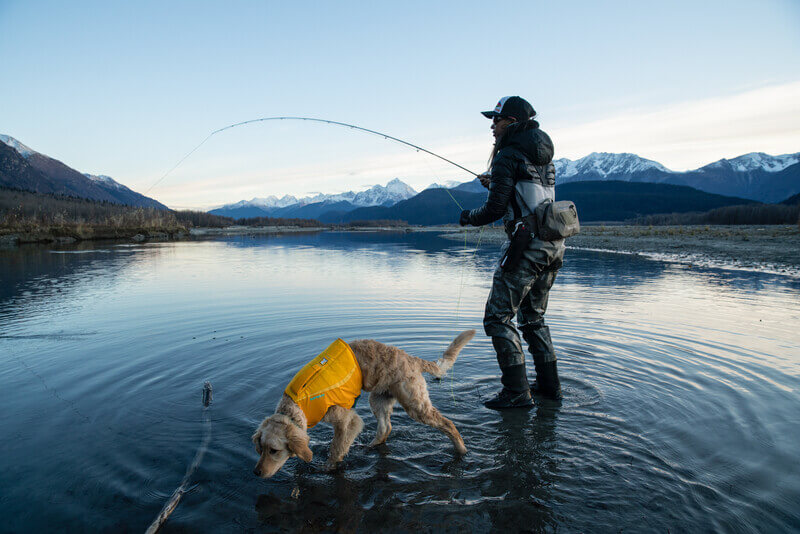 Dog wearing dog life jacket in water next to person fishing